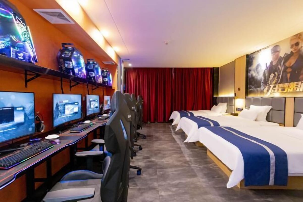 A hotel room with a gaming setup, including multiple monitors, gaming chairs, and a large bed with a blue and white striped bedspread.