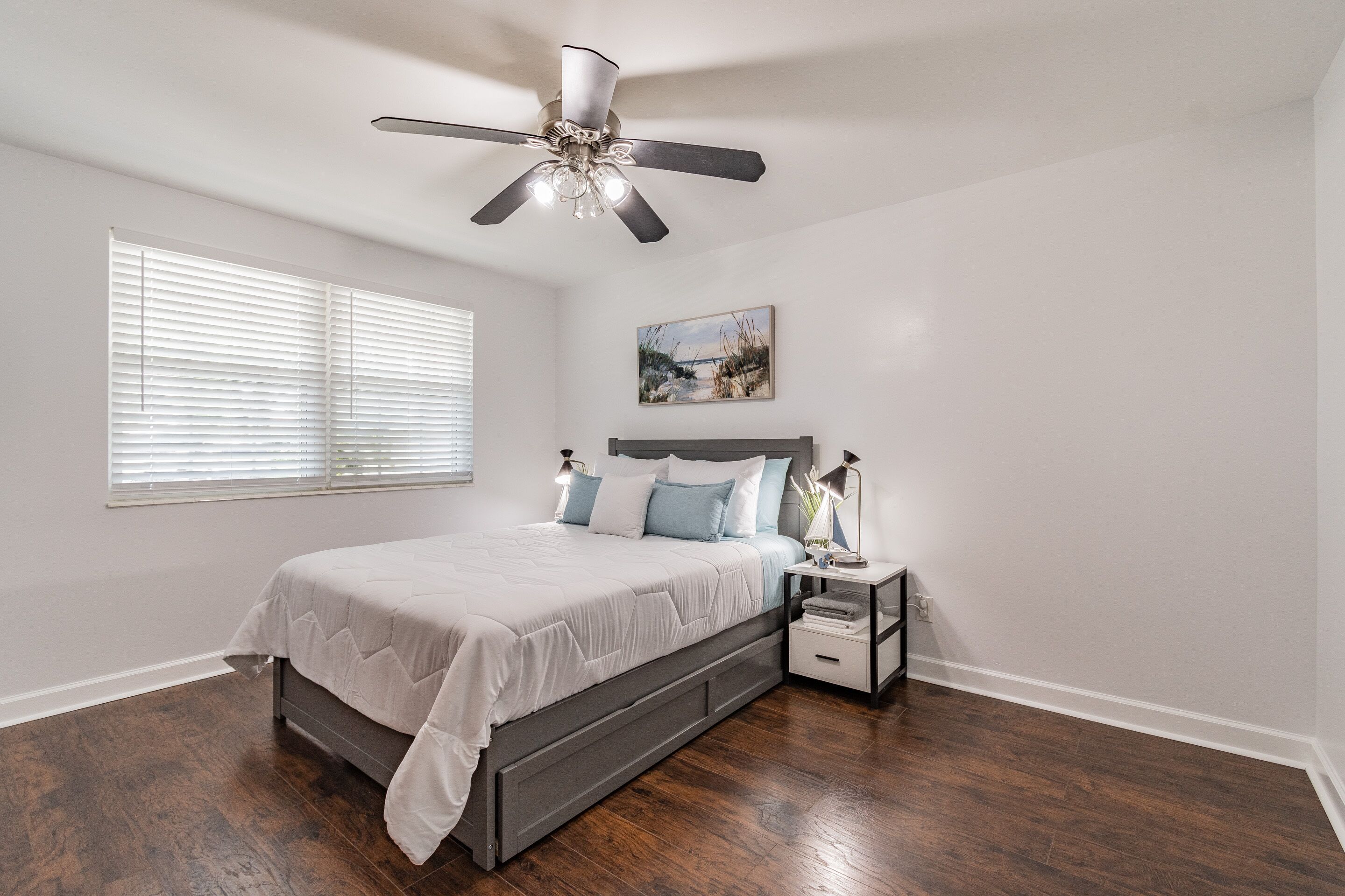 A bedroom with a bed, nightstands, a ceiling fan, a painting, and a window with blinds.