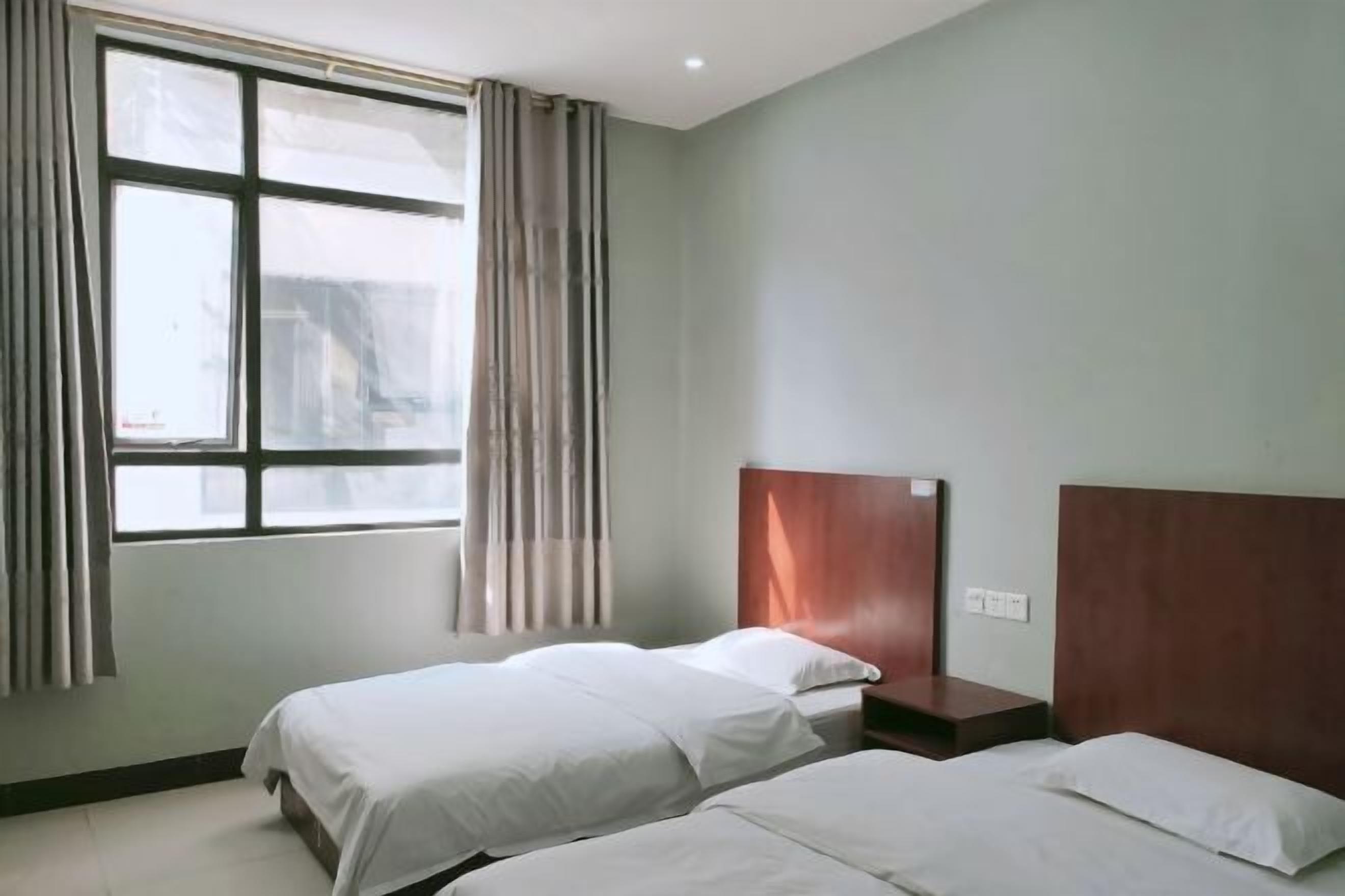 A hotel room with two beds, a wooden headboard, a window with curtains, and a light-colored wall.