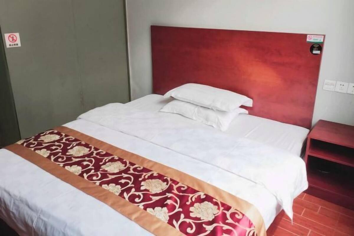 Une chambre d’hôtel avec un lit, une tête de lit rouge, une couvre-lit à motifs et une petite table de chevet en bois.