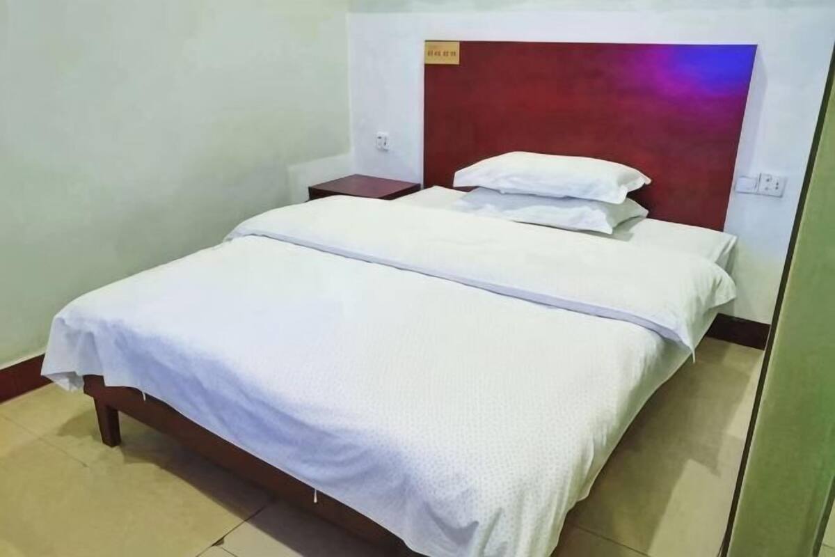 Un lit simple avec une couvre-lit et des oreillers blancs, une tête de lit en bois et une table de chevet avec une lampe.