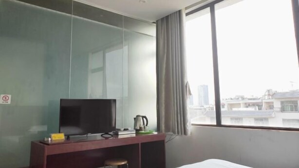 Een hotelkamer met een bed, een bureau met een televisie, een waterkoker en uitzicht op gebouwen door een groot raam.