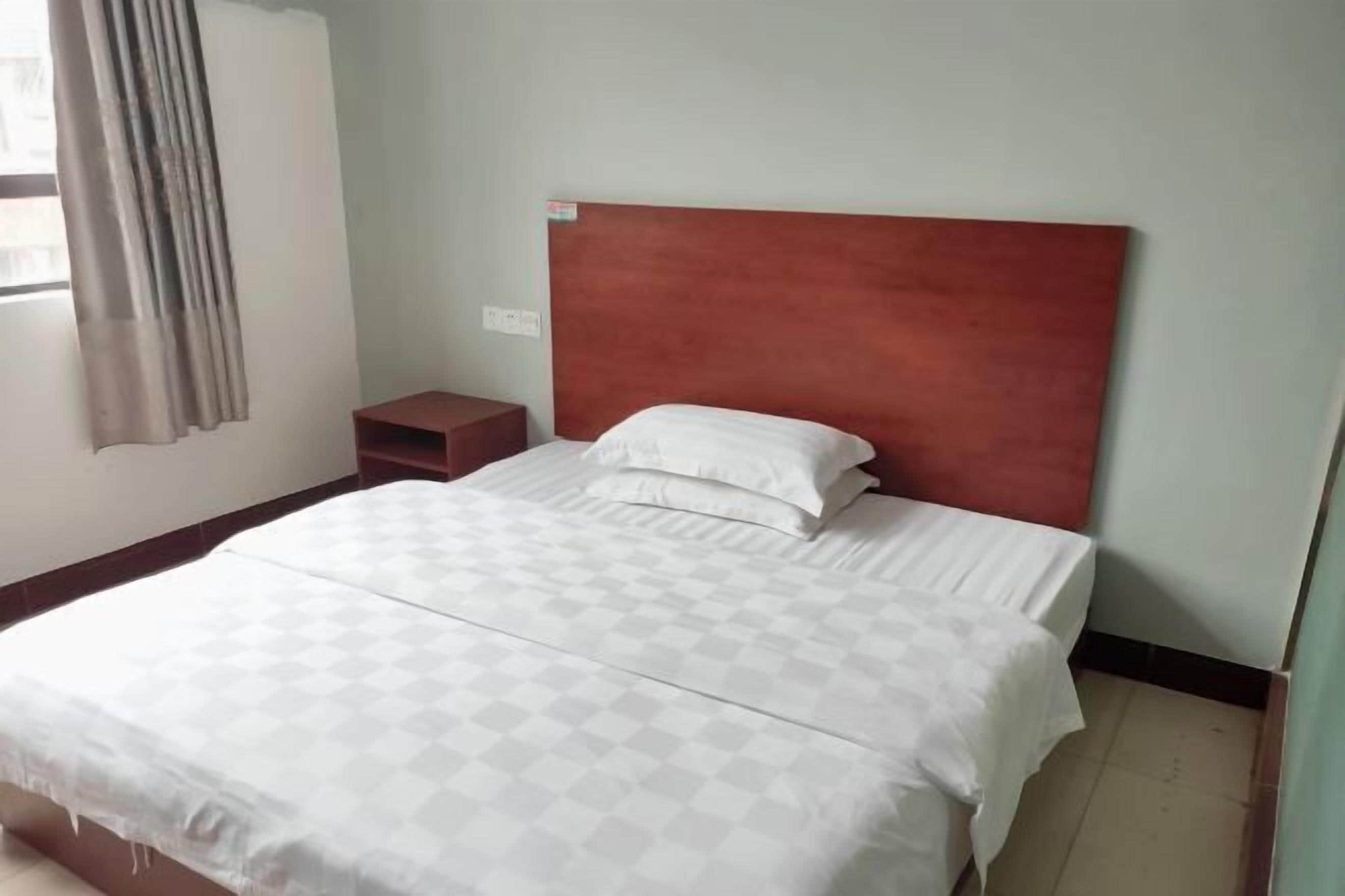 A hotel room with a bed, a nightstand, a window with curtains, and a wall socket.