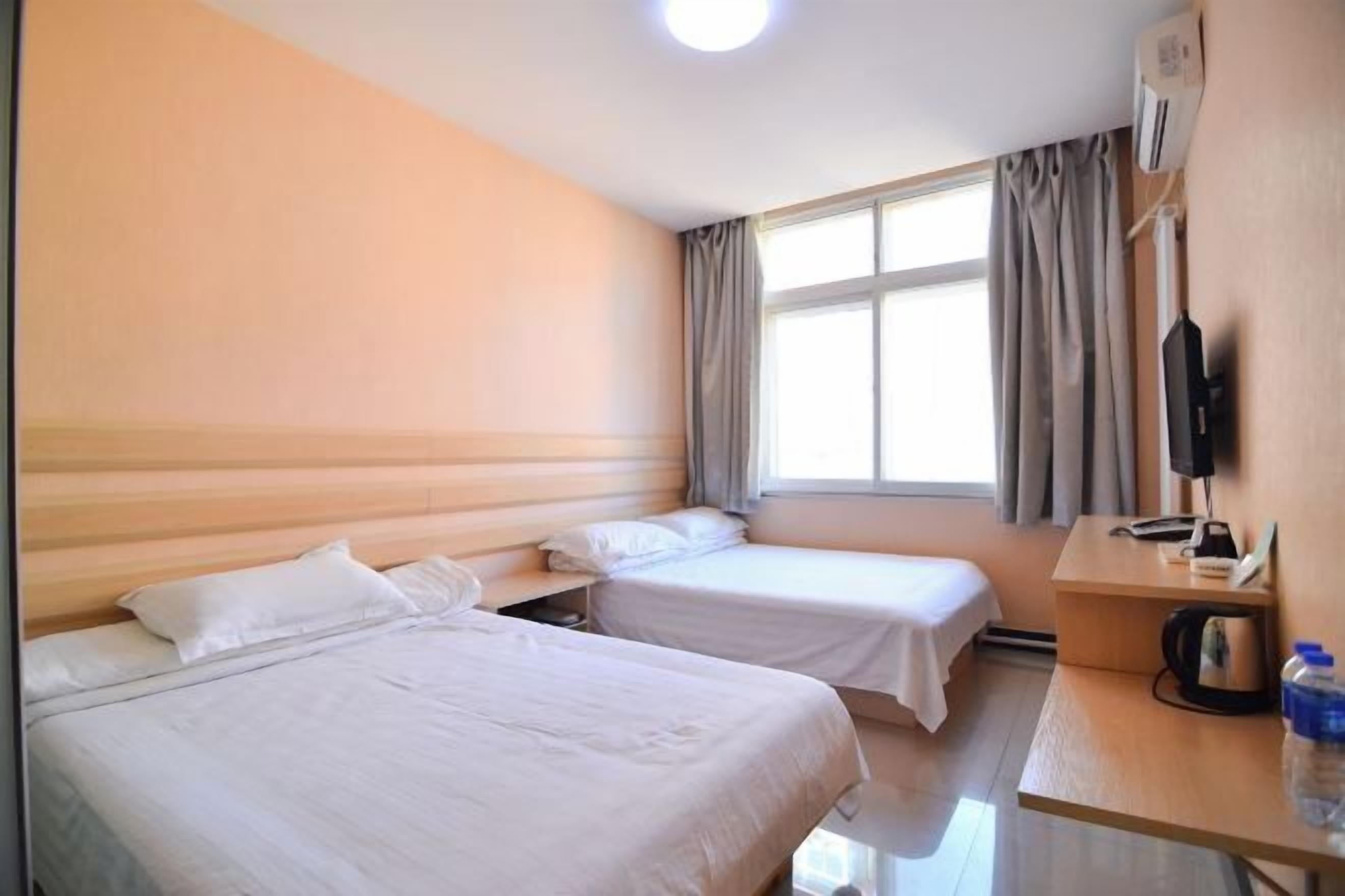 A hotel room with two beds, a desk with a chair, a window with curtains, and a flat-screen TV.