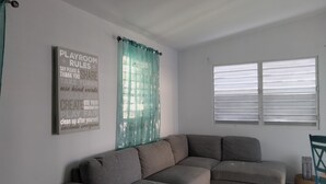 Una sala moderna con un sofá seccional gris, un cartel de normas para la sala de juegos, una ventana con persianas y una cortina verde azulado.