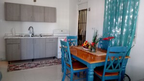 Un comedor con mesa de madera y sillas azules, una cocina con armarios grises y un ventanal con reja de metal.