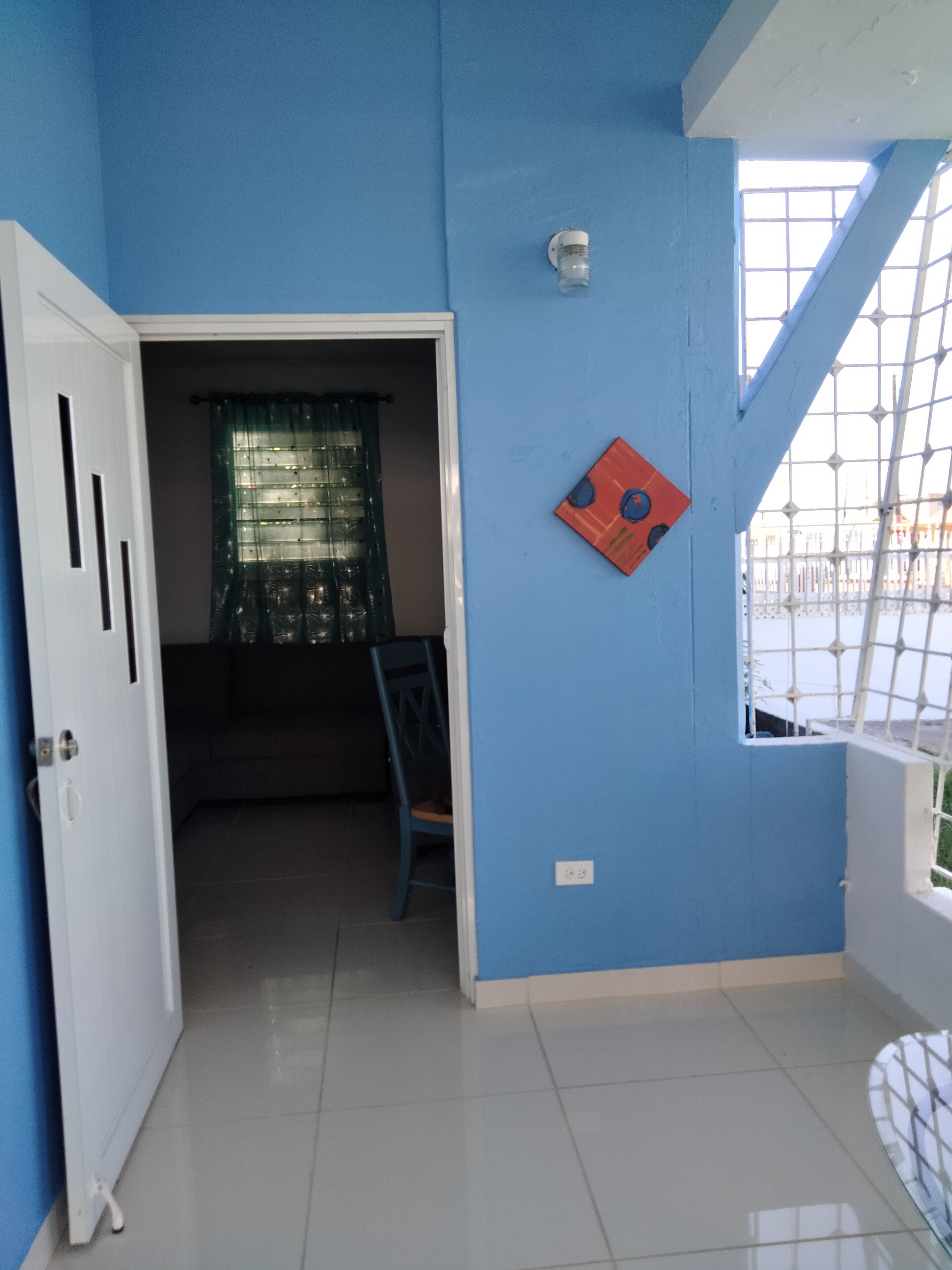 Ein Zimmer mit blauen Wänden, einer weißen Tür, einem Fenster mit Metallgitter und einem Stuhl.