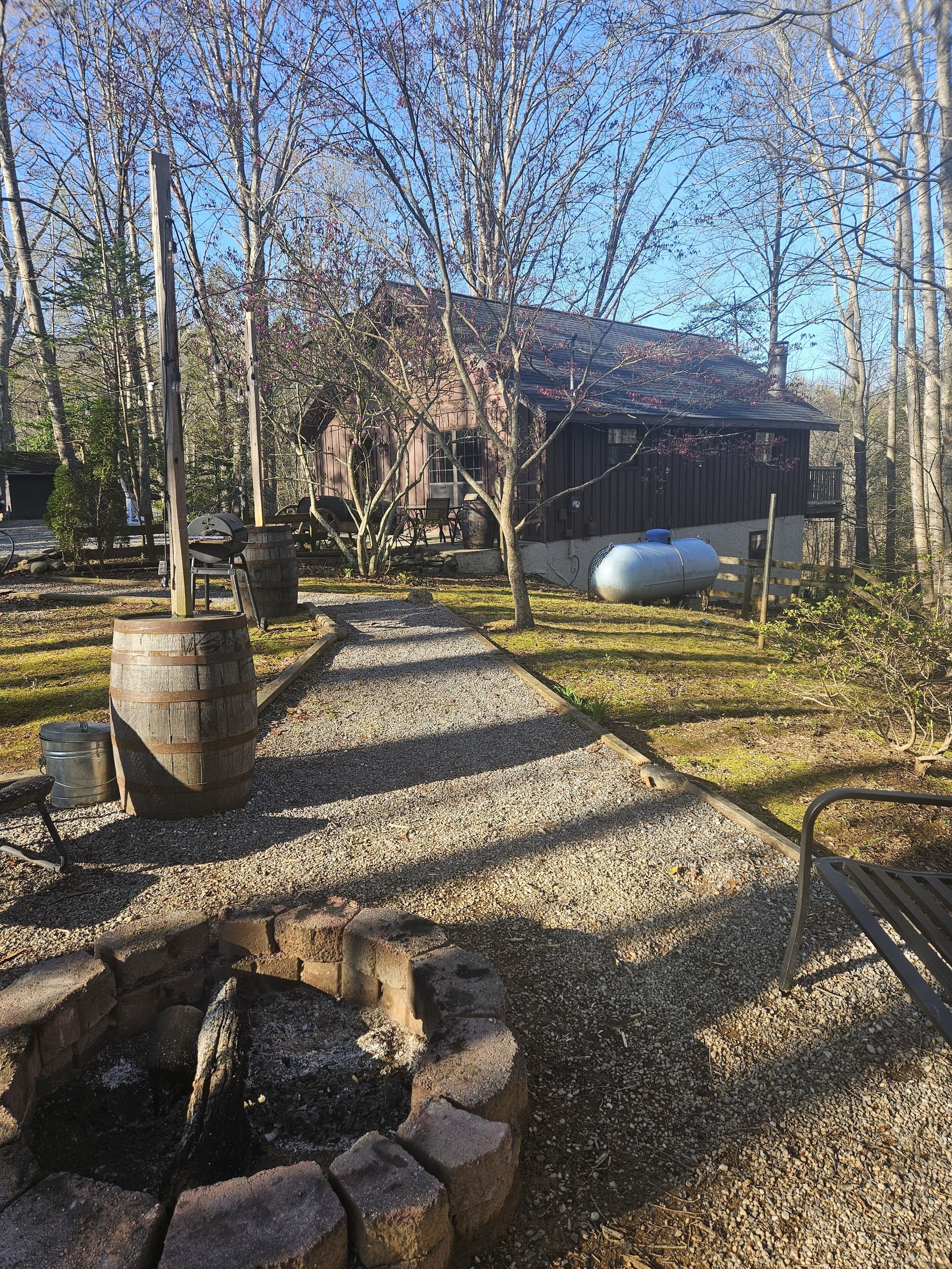 Un espace extérieur rustique agrémenté d’un chemin de gravier, de barils en bois, d’un baril en métal et d’un foyer en pierre.