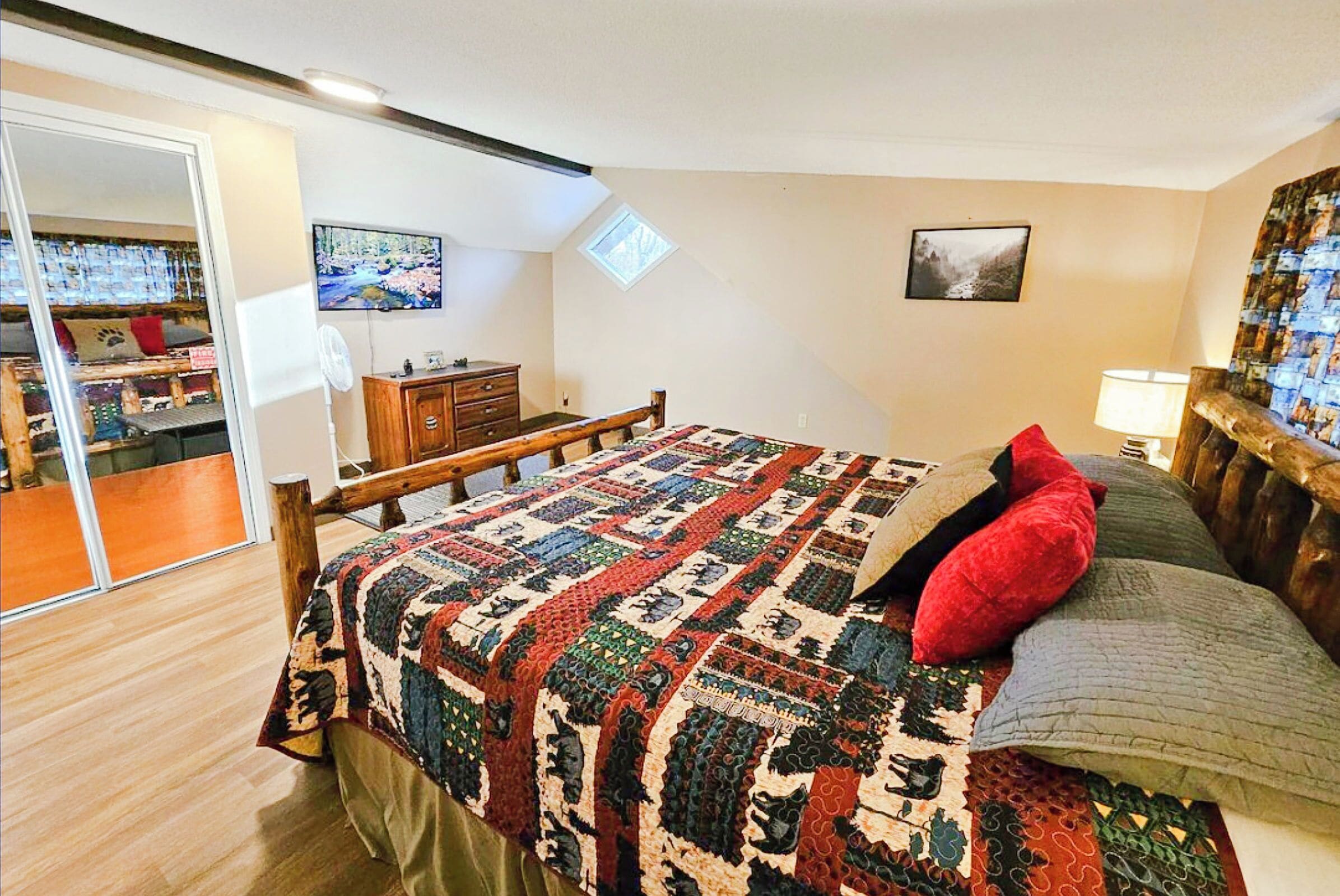 Un dormitorio con cama de madera, colcha, almohada roja, mesita de noche, lámpara y ventana con vistas a la montaña.