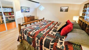 Un dormitorio con cama de madera, colcha, almohada roja, mesita de noche, lámpara y ventana con vistas a la montaña.