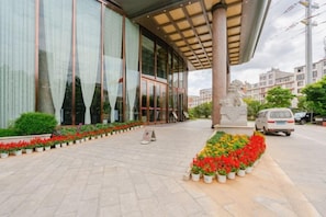 Edifício com amplas janelas, uma estátua e uma área de entrada pavimentada com canteiros de flores.