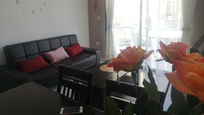 A living room with a black sofa, red and pink cushions, a wooden chair, a glass table, and a smartphone on it.