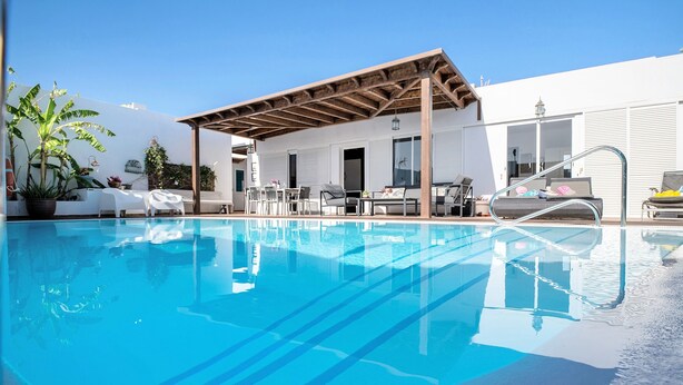 A swimming pool with a covered area, lounge chairs, and a patio with potted plants.