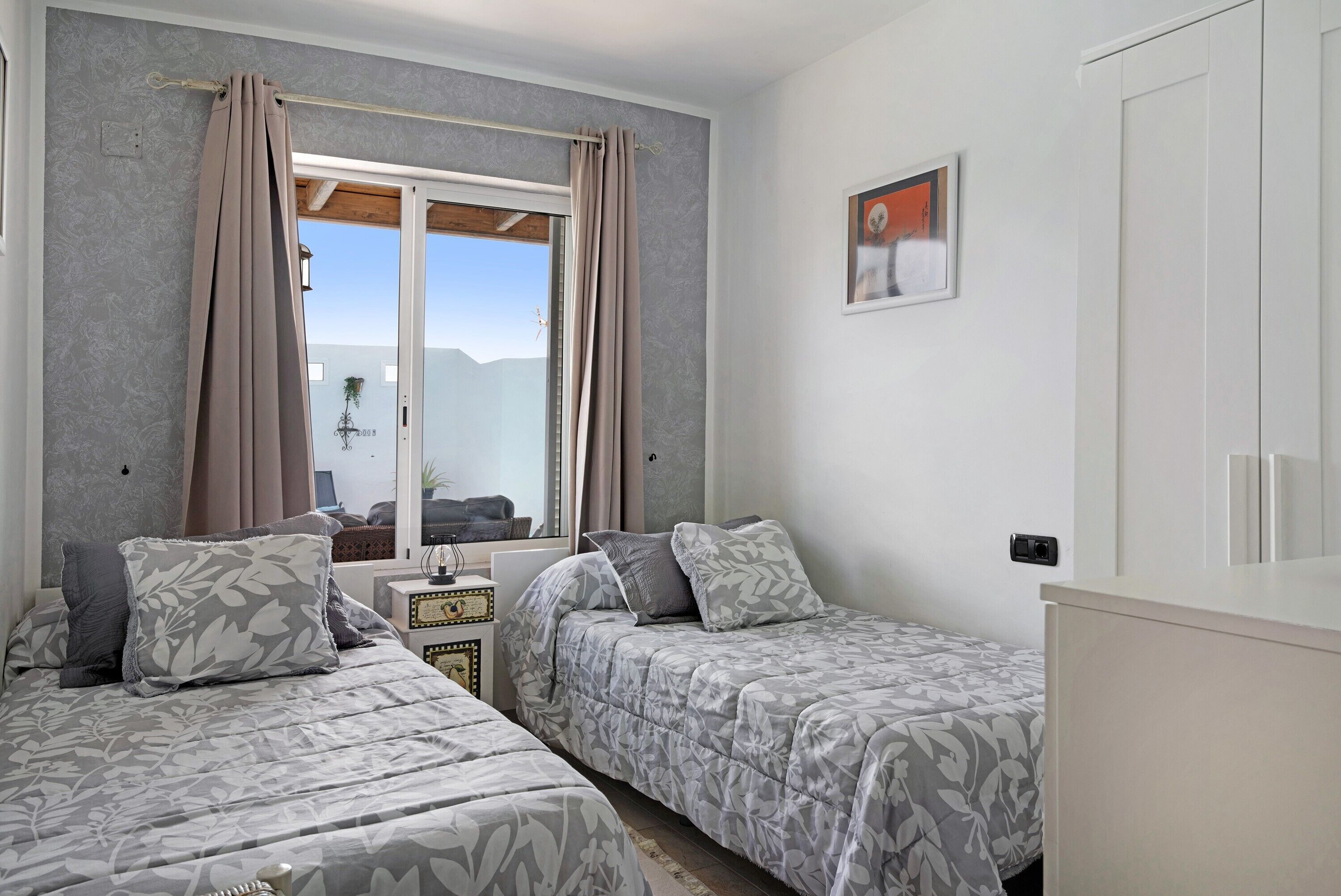 A bedroom with two beds, a window with curtains, a small table, and a wall-mounted picture.