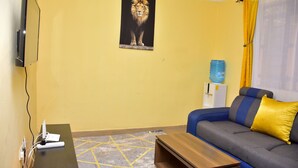 Een woonkamer met een bank, een salontafel, een wandgemonteerde televisie, een waterdispenser en een poster aan de muur.