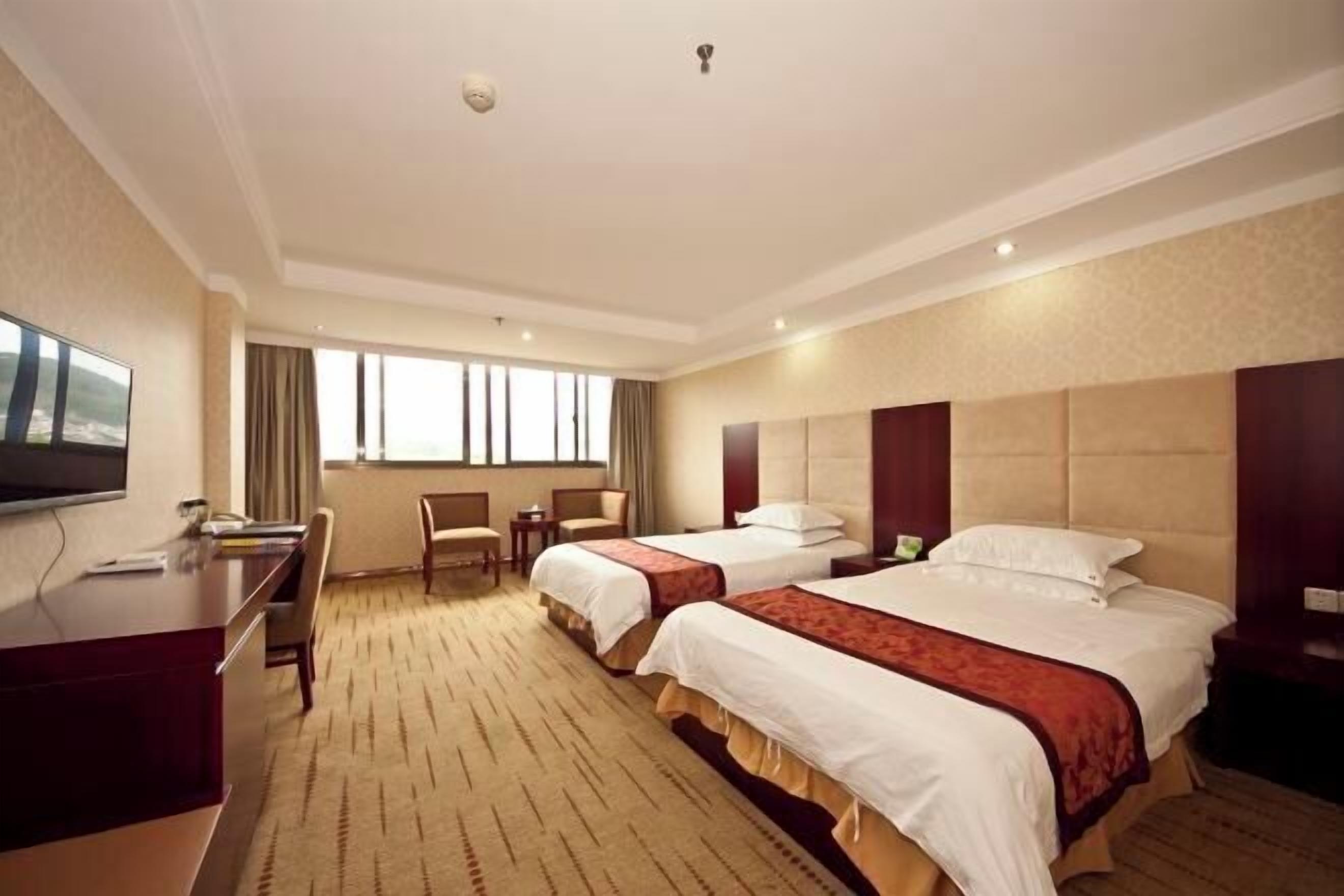 A hotel room with two beds, a desk, a TV, and a large window.