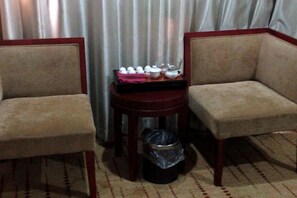 Una habitación con dos sillones color beige, una mesita de madera con una bandeja de tazas y platillos blancos, y un cubo de basura.