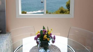 A dining area with a table and chairs, a window with a view of the sea and boats, and a vase with flowers on the table. - Dream Luxury Beach Front Majestic Sea-views (Palmanova)