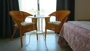 A room with two wicker chairs, a small round table, a bed with a patterned bedspread, and a view through a window with curtains. - Dream Luxury Beach Front Majestic Sea-views (Palmanova)