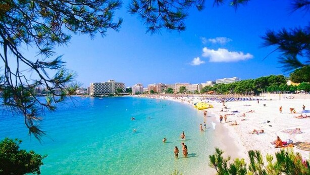 A beach with clear turquoise water, sandy shore, and buildings in the background. - Dream Luxury Beach Front Majestic Sea-views (Palmanova)