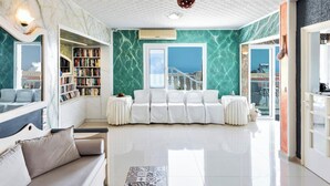 Ampla sala de estar com um sofá branco, uma estante de livros e vista para o mar através de grandes janelas.
