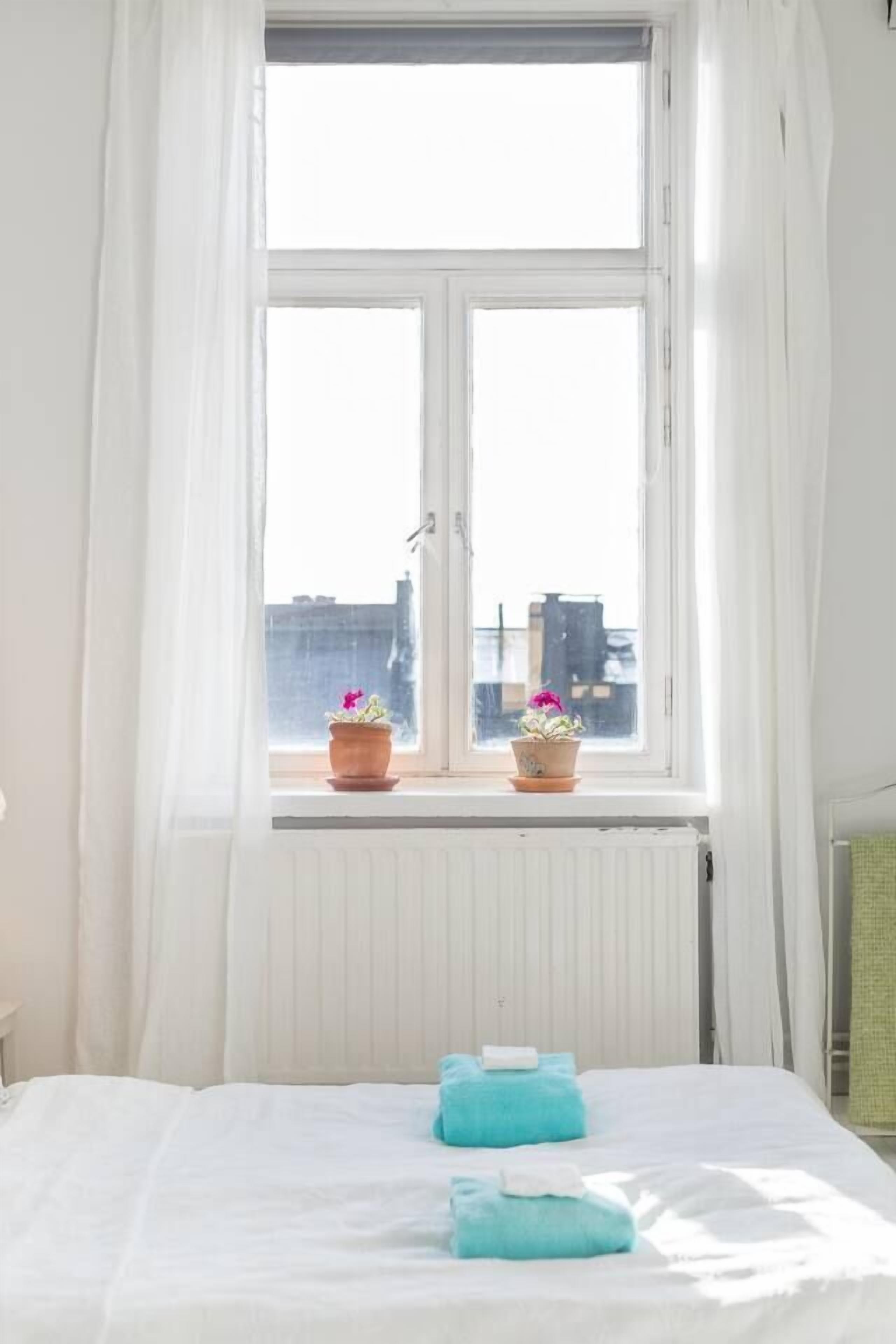 A room with a bed, white curtains, and two potted plants on the windowsill.