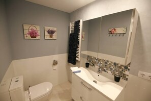 A bathroom with a toilet, sink, and mirror. There are two wall-mounted paintings and a towel rack. - Happy Luxury Best Beach Majestic Sea-Views (Palmanova)