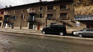 A multi-story building with balconies, a parked black van, and a car on the street. - Apartment by the river with snow peak views (Modane)