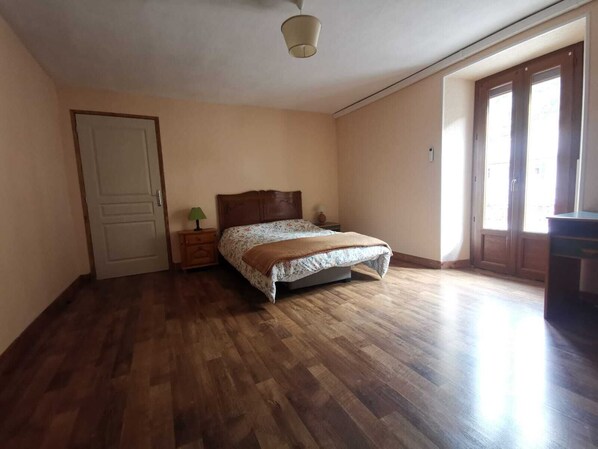 A bedroom with a wooden floor, a bed with a floral bedspread, a nightstand with a lamp, and a door. - Apartment by the river with snow peak views (Modane)
