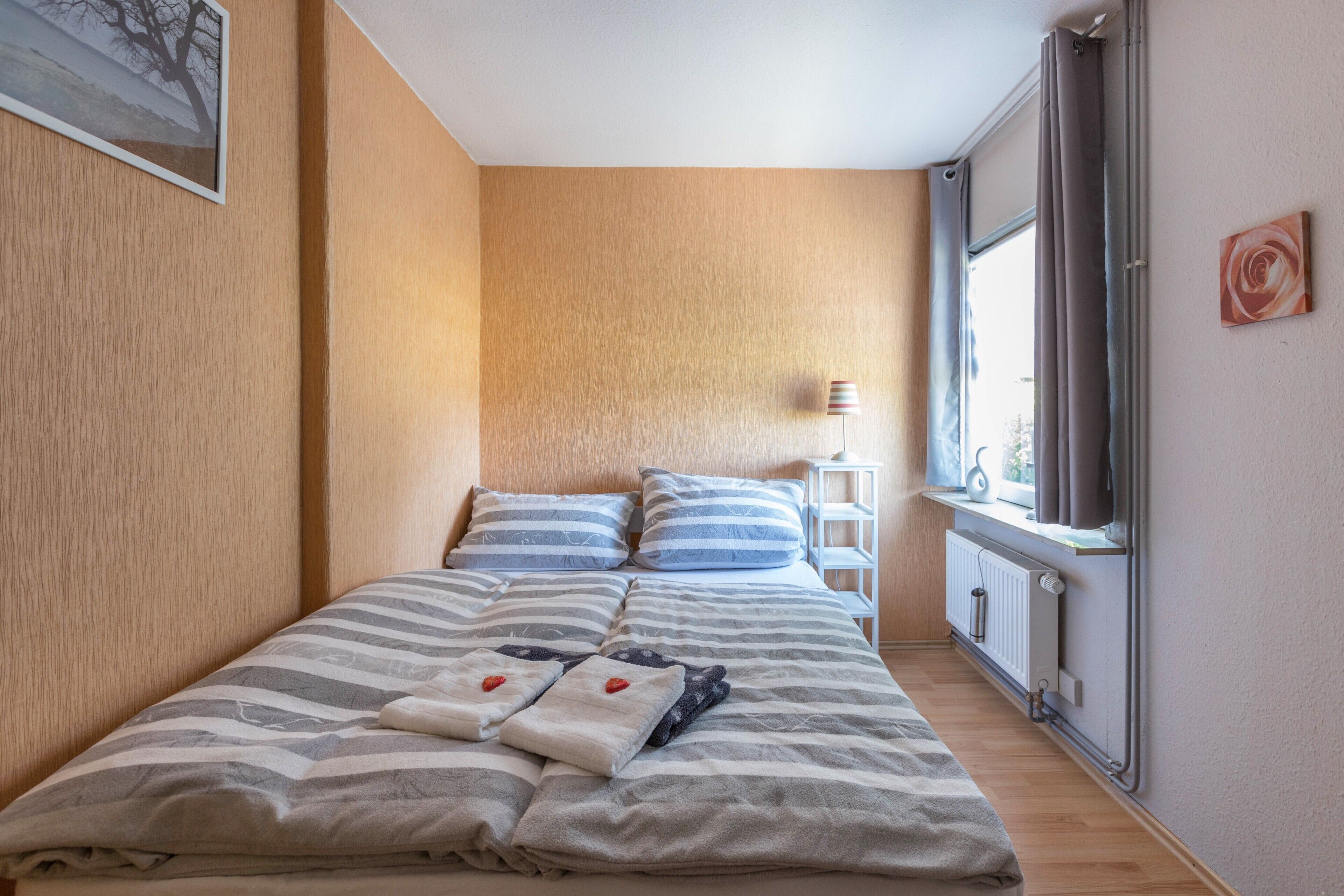 Un dormitorio con cama, almohadas, mesita de noche, lámpara, ventana y radiador.