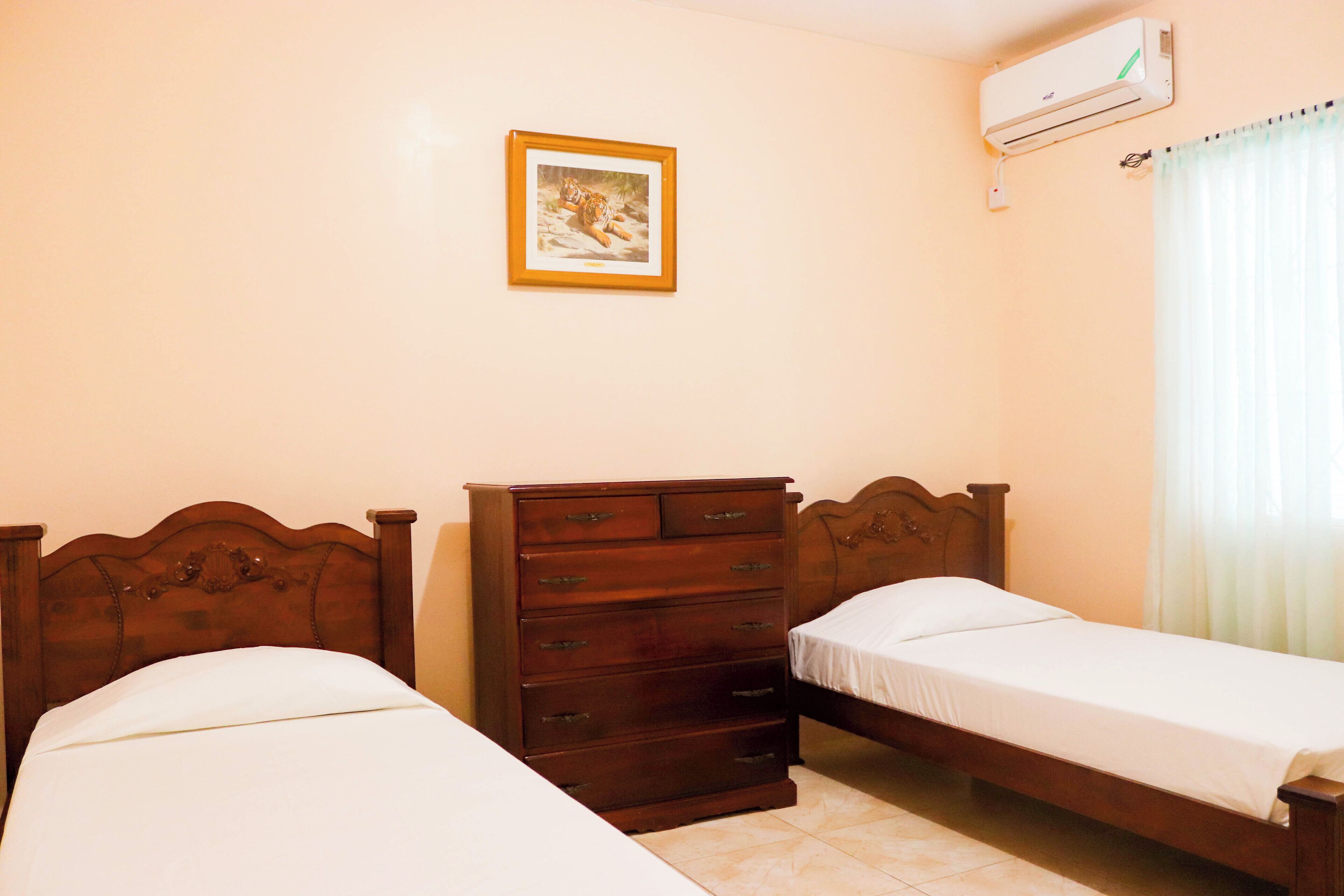A bedroom with two single beds, a wooden dresser, a framed picture on the wall, and an air conditioning unit.