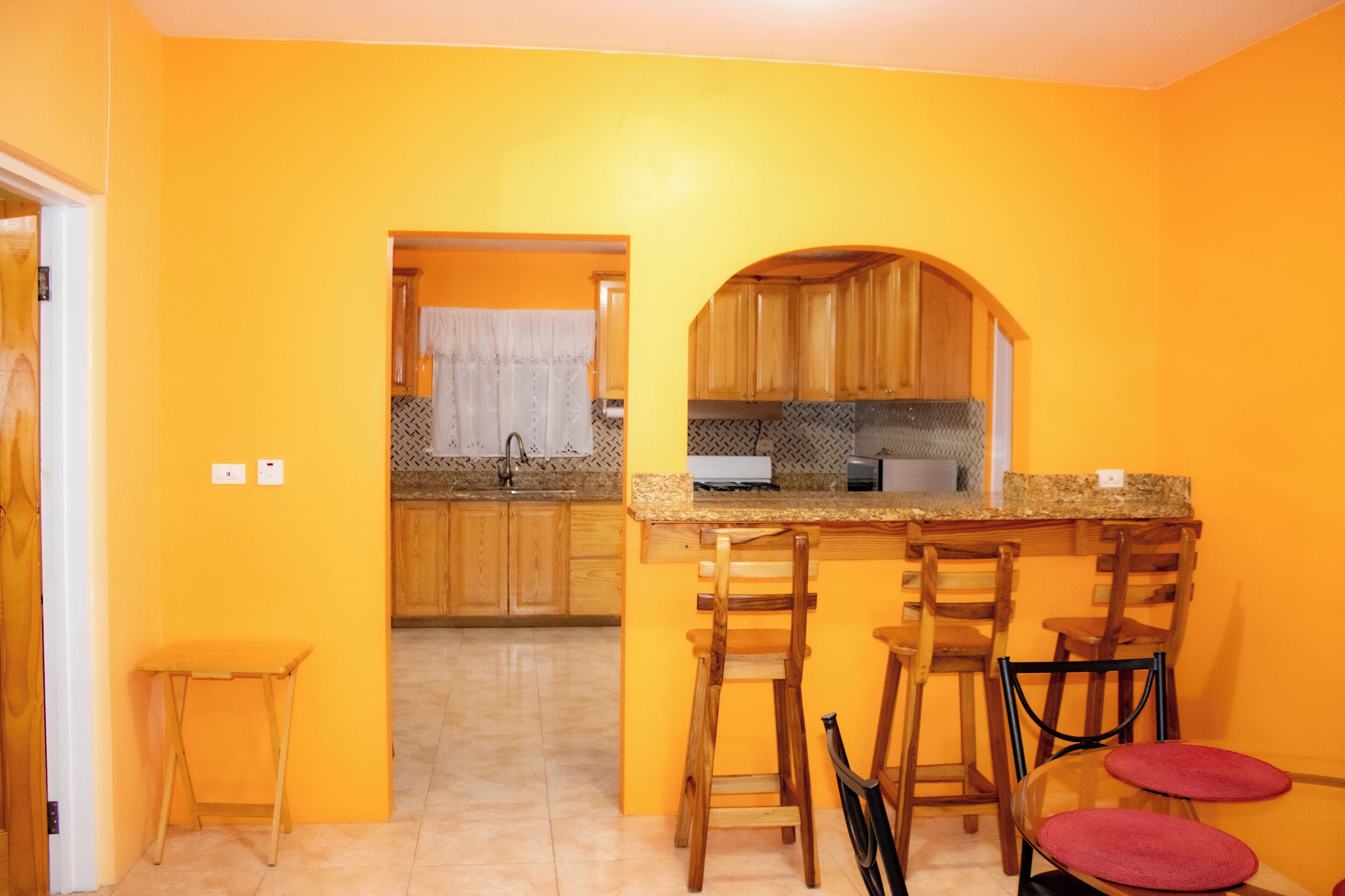 A kitchen with a yellow wall, wooden cabinets, a granite countertop, and a sink. There are three wooden stools with red cushions.