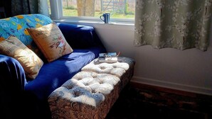 Ein gemütliches Zimmer mit einem blauen Sofa, gemusterten Kissen und Blick auf einen Baum und ein Feld durch ein Fenster.