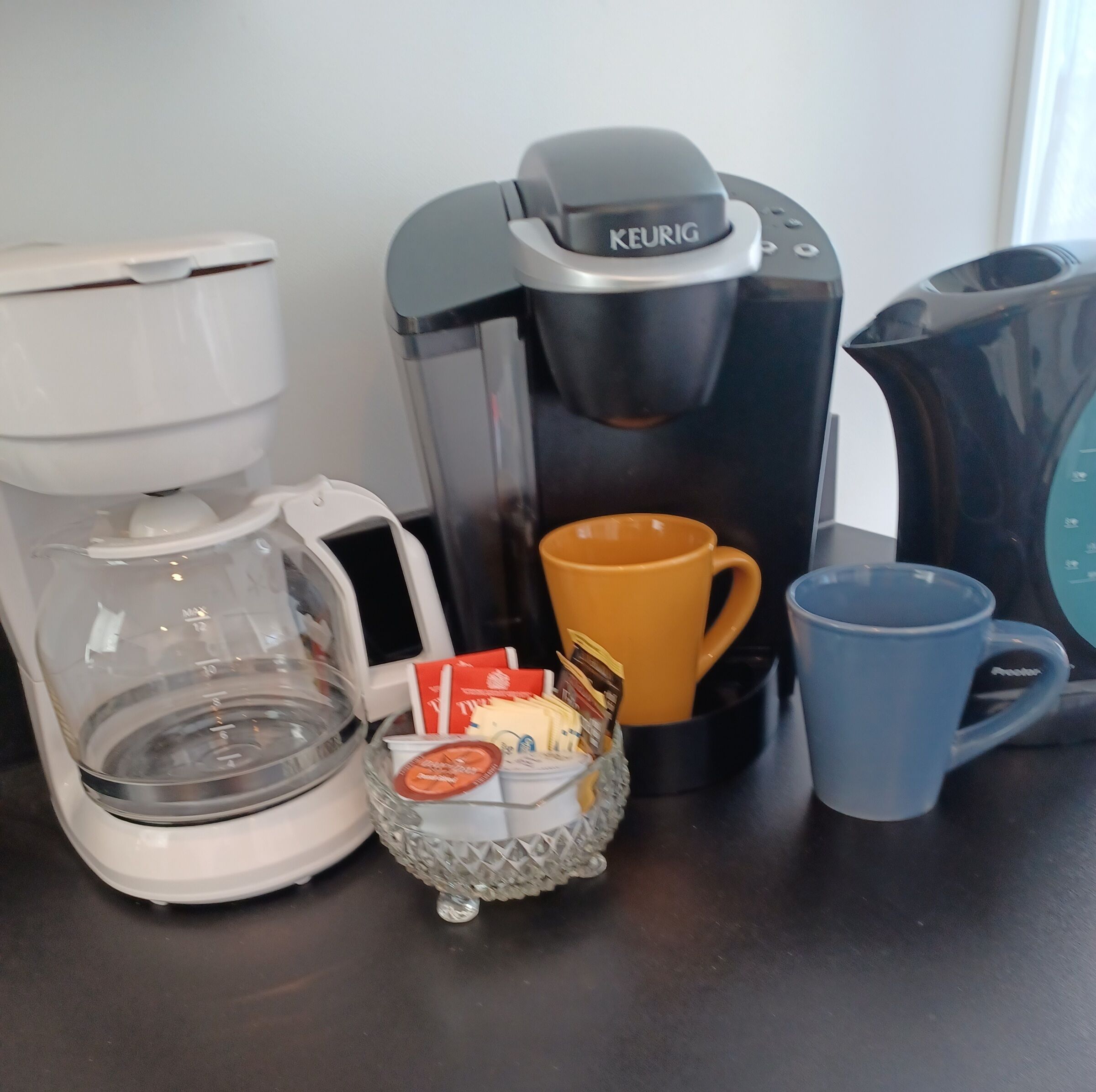 Uma cafeteira, uma jarra de vidro, uma caneca e uma pequena cesta com cápsulas de café em uma bancada.