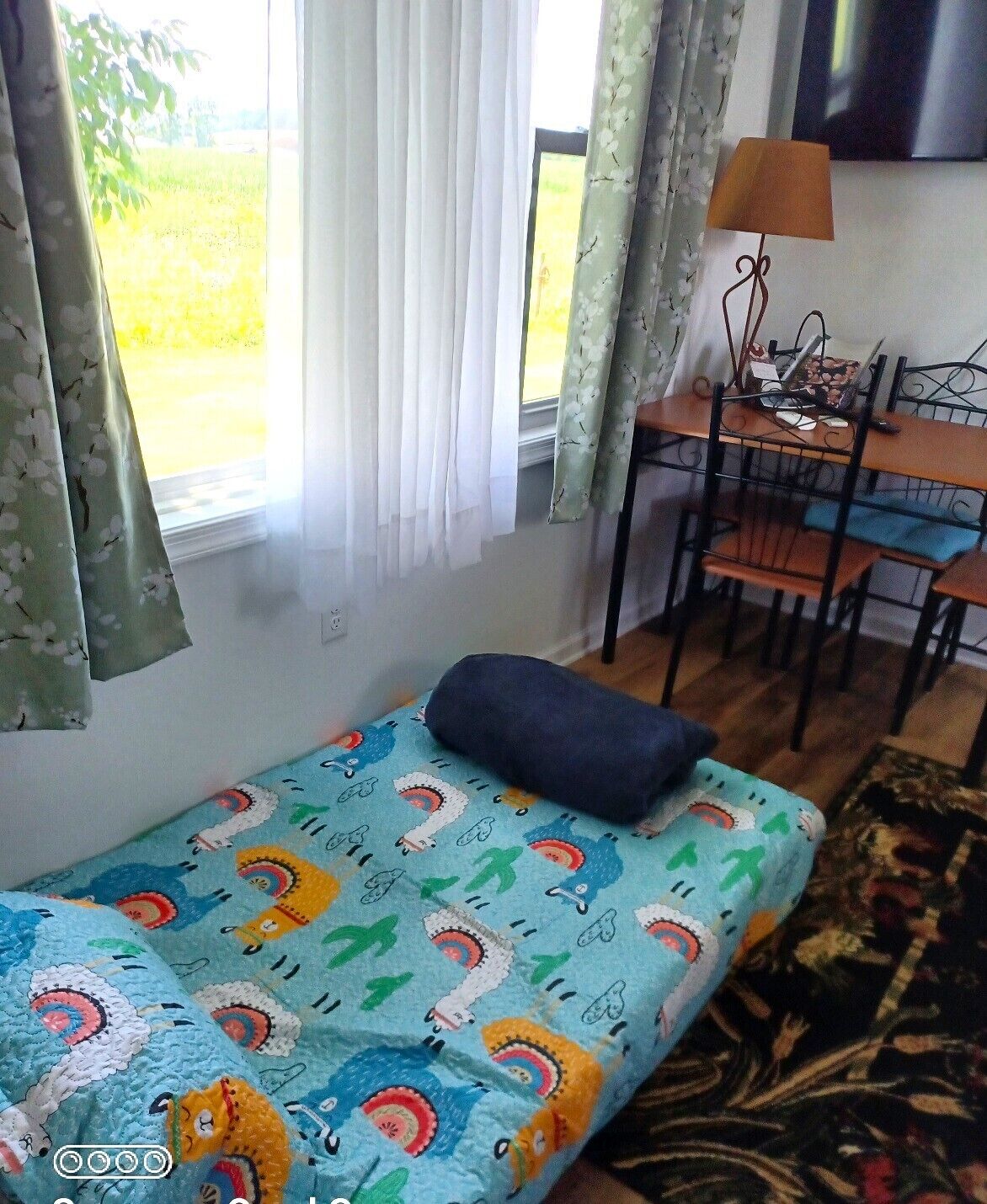 Ein kleines Zimmer mit einem Bett, einem Schreibtisch und einem Stuhl. Ein Fenster mit Vorhängen und eine Lampe auf dem Schreibtisch sind ebenfalls vorhanden.