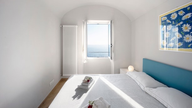 A bedroom with a bed, a window, a radiator, and a wall with a floral pattern. - Vacation Apartment in the Historic Center with Sea View and Shared Garden (Bordighera)