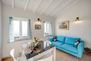 A room with a blue sofa, a dining table with chairs, a vase with flowers, and two windows with curtains.