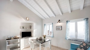A modern living room with a dining area, white furniture, a flat-screen TV, and a sofa with a floral cushion. - Vacation Apartment in the Historic Center with Sea View and Shared Garden (Bordighera)