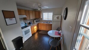 A compact kitchen with wooden cabinets, a microwave, a stove, and a small dining area with a table and chairs. - Furnished house in Geraldton (Geraldton)