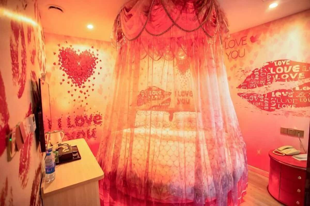 Una habitación con una cama cubierta por una tela ligera roja, una pared con adornos con forma de corazón y un pequeño escritorio con una botella de agua.