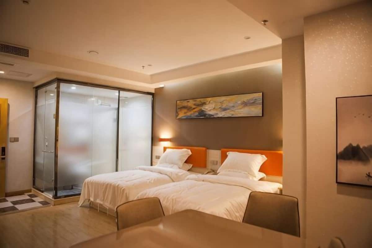 Habitación de hotel con dos camas, cabina de ducha de vidrio, mesa de comedor y obras de arte en las paredes.