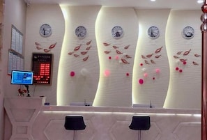 A reception area with a modern design, featuring a curved wall with clocks and decorative elements. There is a digital display board and a reception desk with chairs.