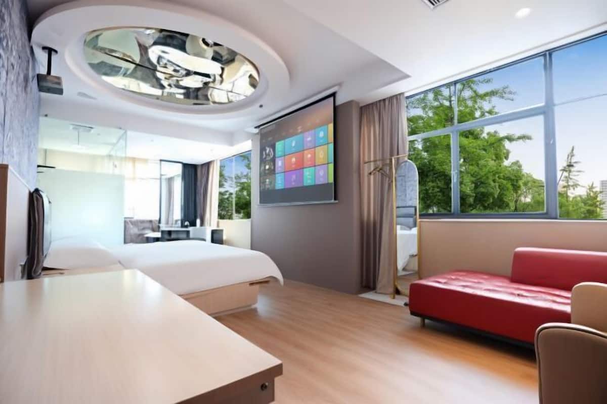 Moderni hotellihuone, jossa on sänky, työpöytä, punainen sohva, suuri ikkuna, josta on näkymä, ja katossa pyöreä kattovalaisin.