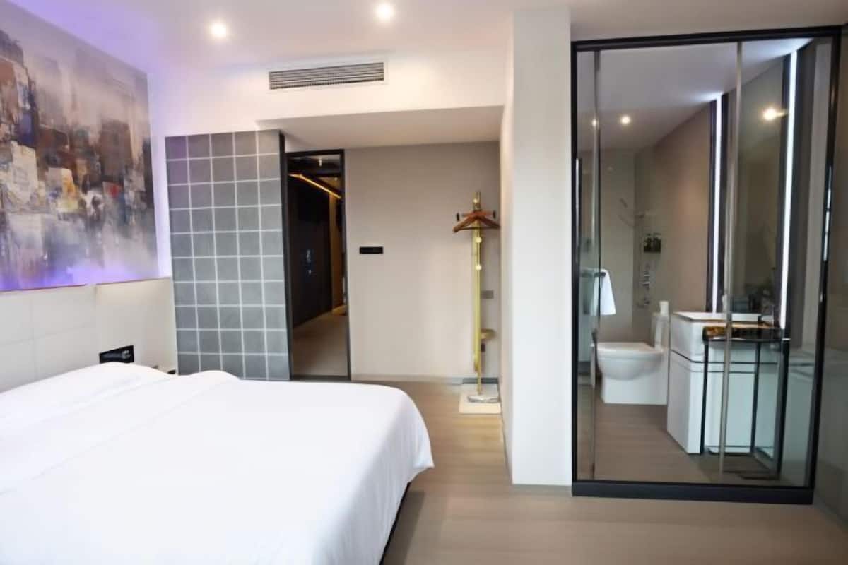 Moderni hotellihuone, jossa on suuri sänky, lasiseinä, kylpyhuone, jossa on wc ja pesuallas, sekä taulu seinällä.