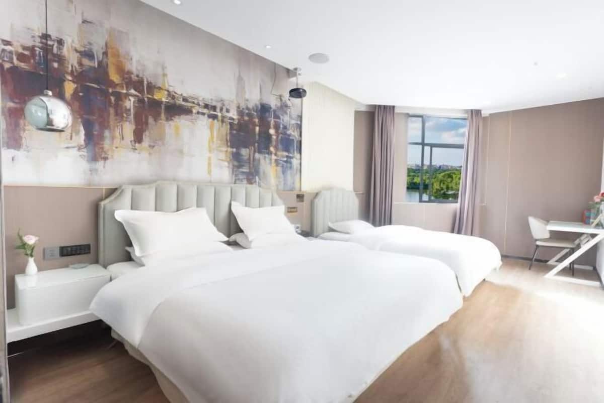 Moderni hotellihuone, jossa on kaksi sänkyä, suuri abstrakti taulu seinällä, työpöytä tuolilla ja näkymä vehreään maisemaan.