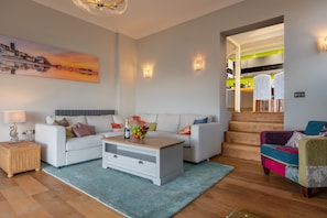 A modern living room with a white sofa, a coffee table, and a colorful armchair. There is a staircase leading to a kitchen area. - Boycott House, Cromer, Norfolk (Cromer)
