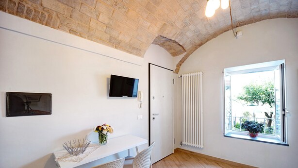 Ein Zimmer mit weißer Wand, einem Fernseher über einem Tisch mit Stühlen, einer Tür, einem Fenster mit Jalousien und einem Heizkörper.