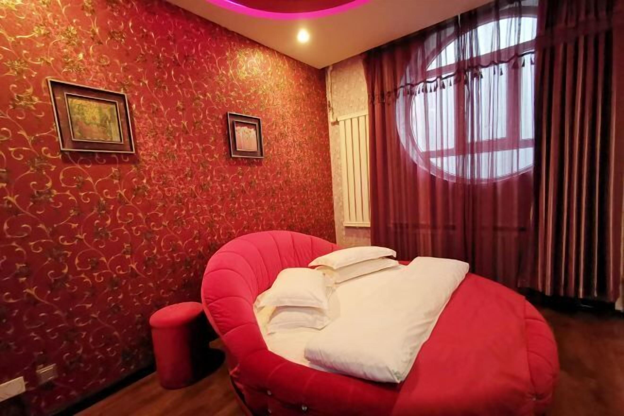 A room with a red patterned wall, a red chair with pillows, a small red stool, and a window with curtains.