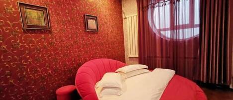 A room with a red patterned wall, a red chair with pillows, a small red stool, and a window with curtains.