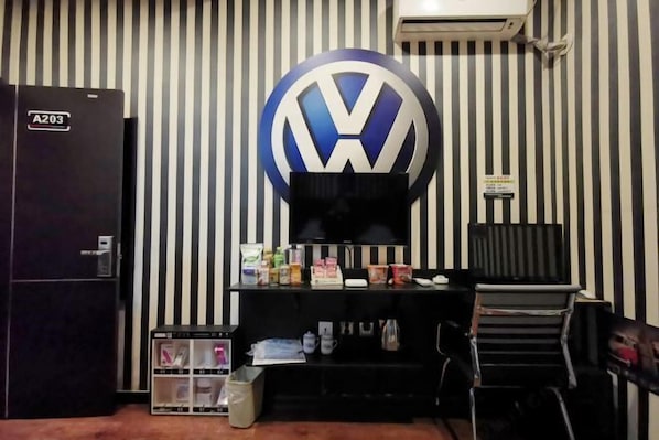 A room with a striped wall, a black A203 refrigerator, a desk with various items, and a VW logo on the wall. - Zhiyoung Theme Hotel (Daqing Xinbaida Dongfeng Middle School) (Daqing)